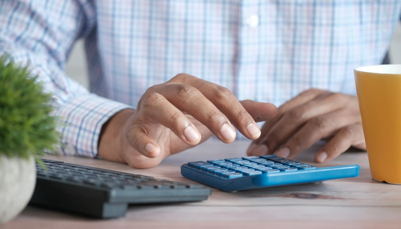 A person using a calculator at a desk beside a keyboard and a mug