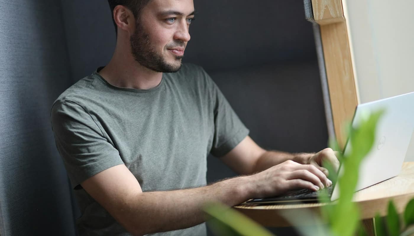 A man smiling as he works on a laptop beside a plant