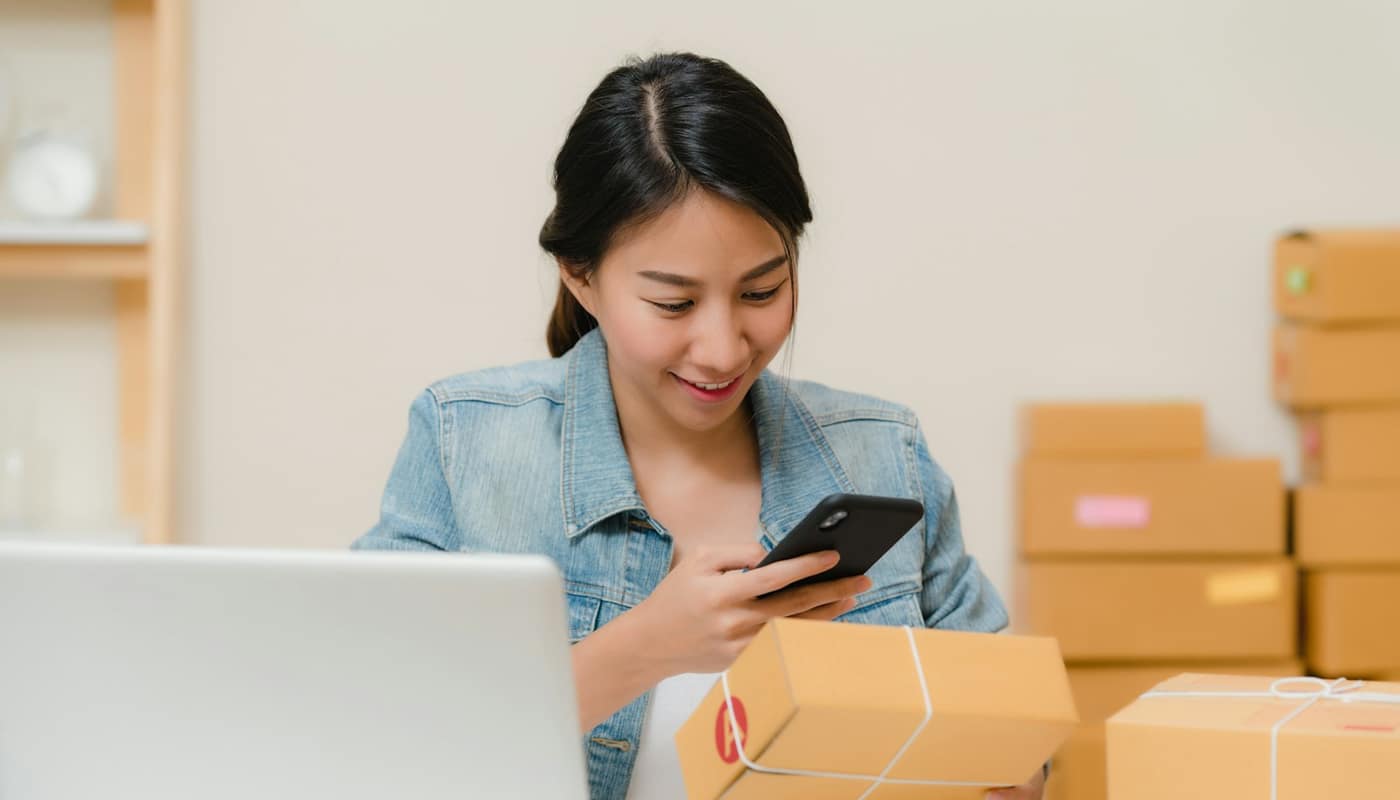 A woman checking a parcel on her phone beside a laptop and stacked cardboard boxes