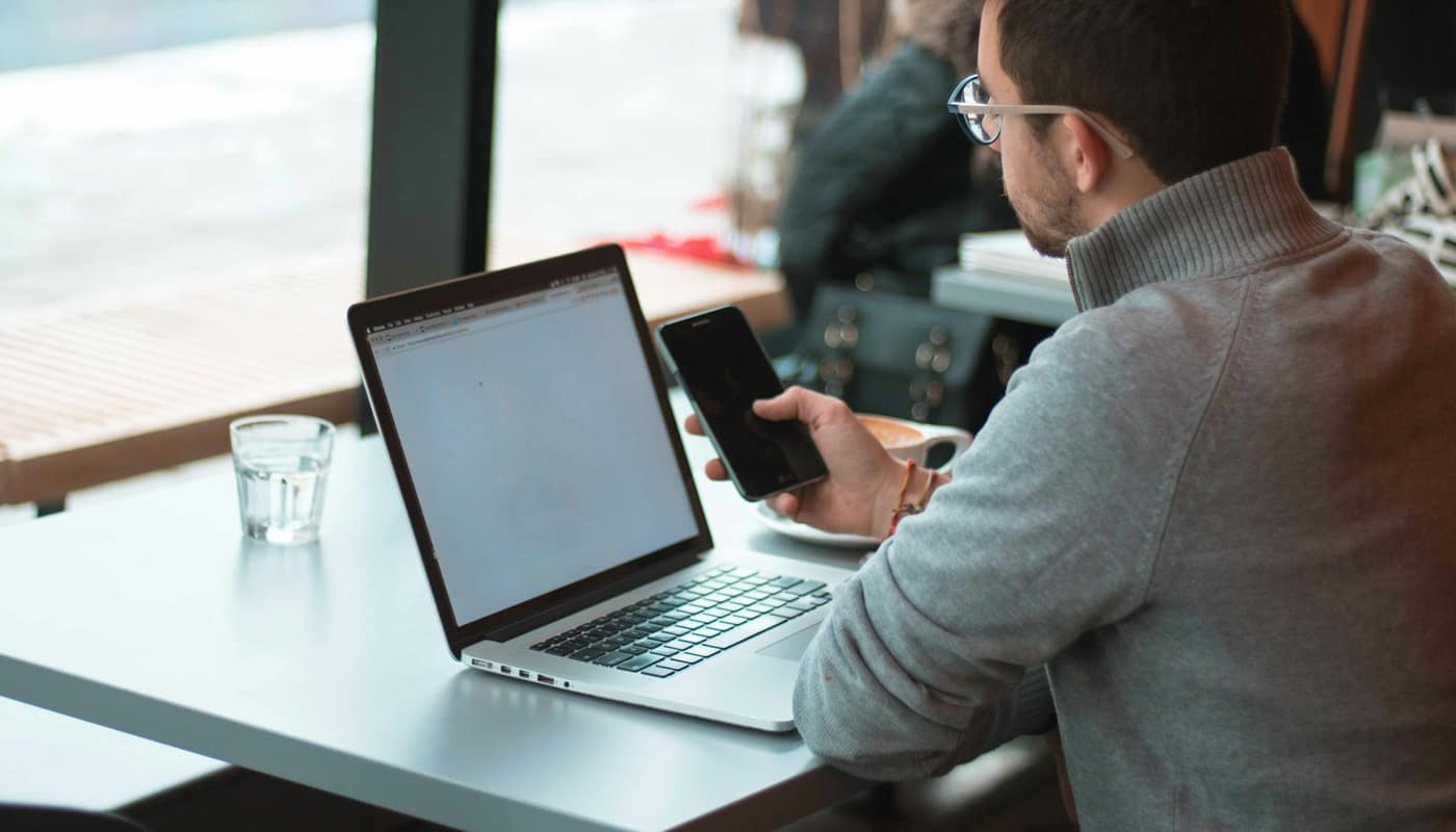 A man working on a laptop at a table by a window, with a phone and a glass of water