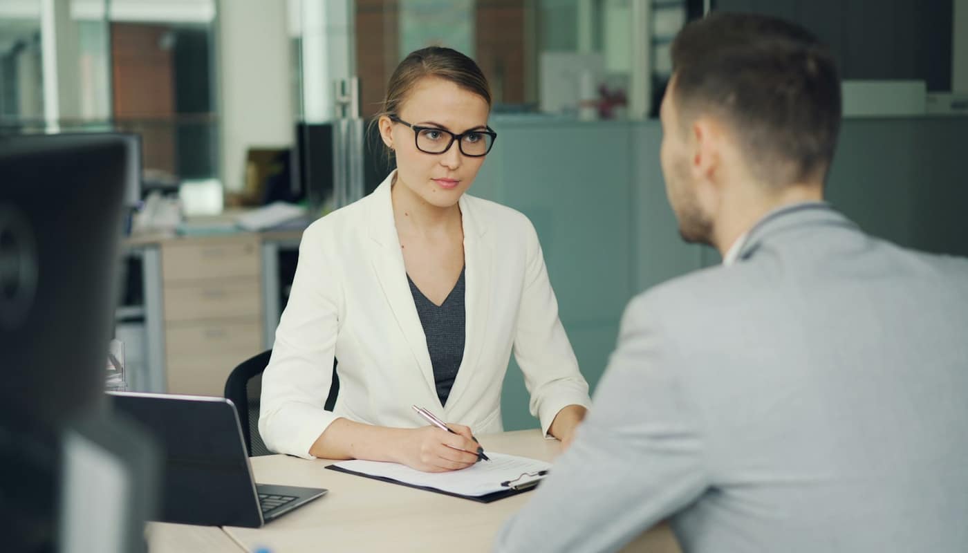 A woman in glasses and a white blazer interviewing a man across a desk