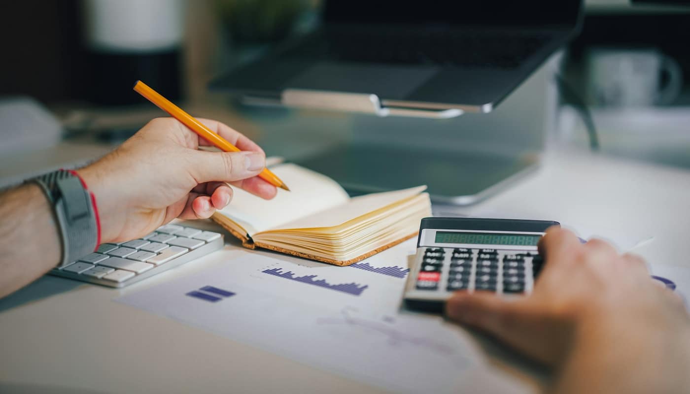 A hand writing in a notebook beside a calculator on a desk