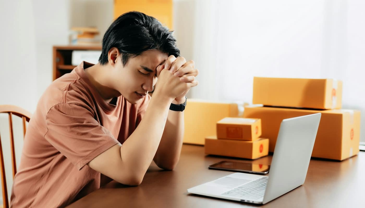 A man resting his head on his hands at a table with a laptop and cardboard boxes