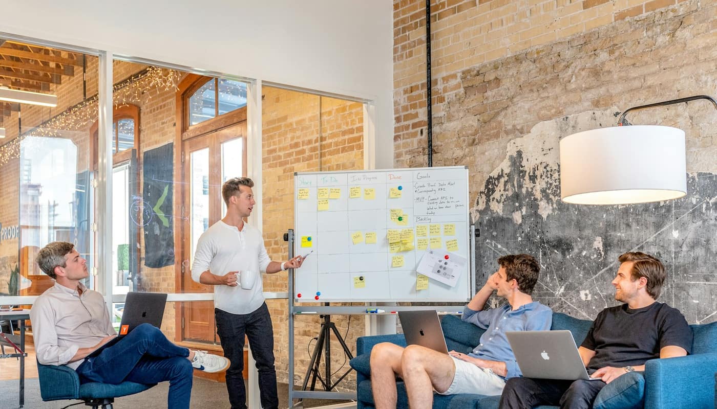 A team on laptops while a colleague stands at a whiteboard in a brick-walled office